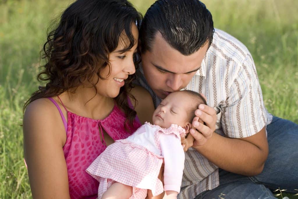 A mother and father adoring their baby outdoors, with the mother in pink and the father in a striped shirt