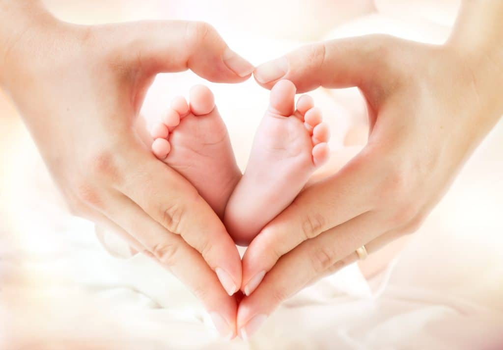 Adult hands forming a heart shape around a baby's bare feet against a soft, warm background